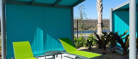 A covered outdoor seating area with two green lounge chairs and a small table, surrounded by a blue fence and plants.