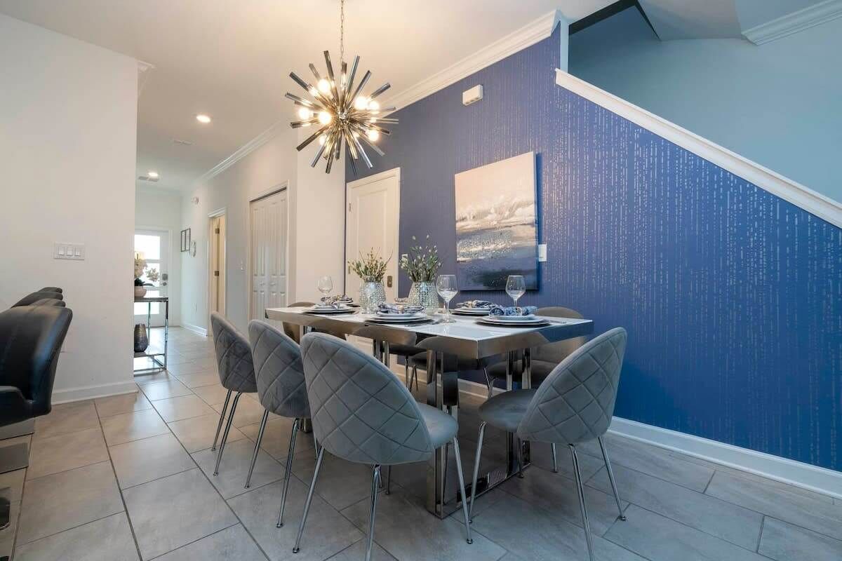 A dining area with a large table set for six, grey upholstered chairs, a modern chandelier, and a blue textured wall.