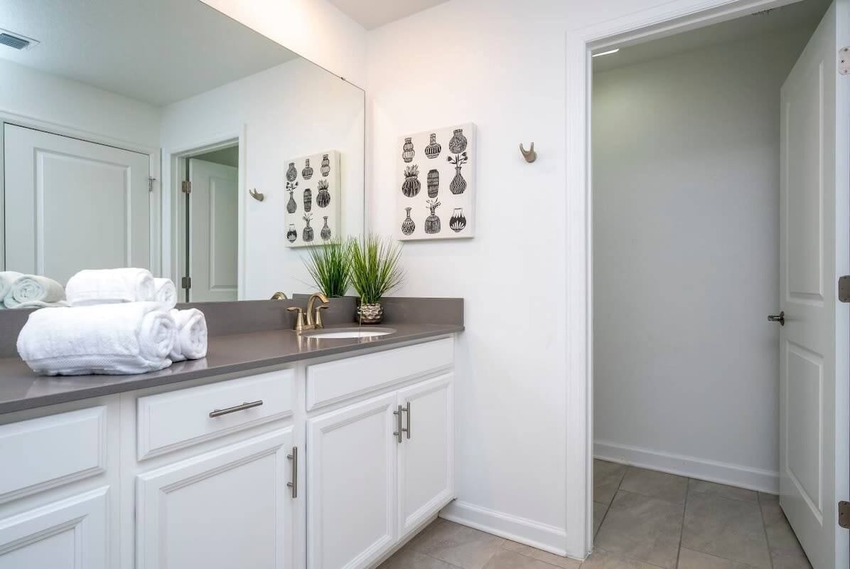 Une salle de bain avec des meubles blancs, un grand miroir et deux œuvres d’art accrochées au mur, représentant des vases.