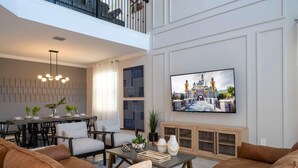 Uma sala de estar espaçosa com teto alto, área de jantar com mesa e cadeiras modernas e uma grande TV de tela plana fixada na parede.