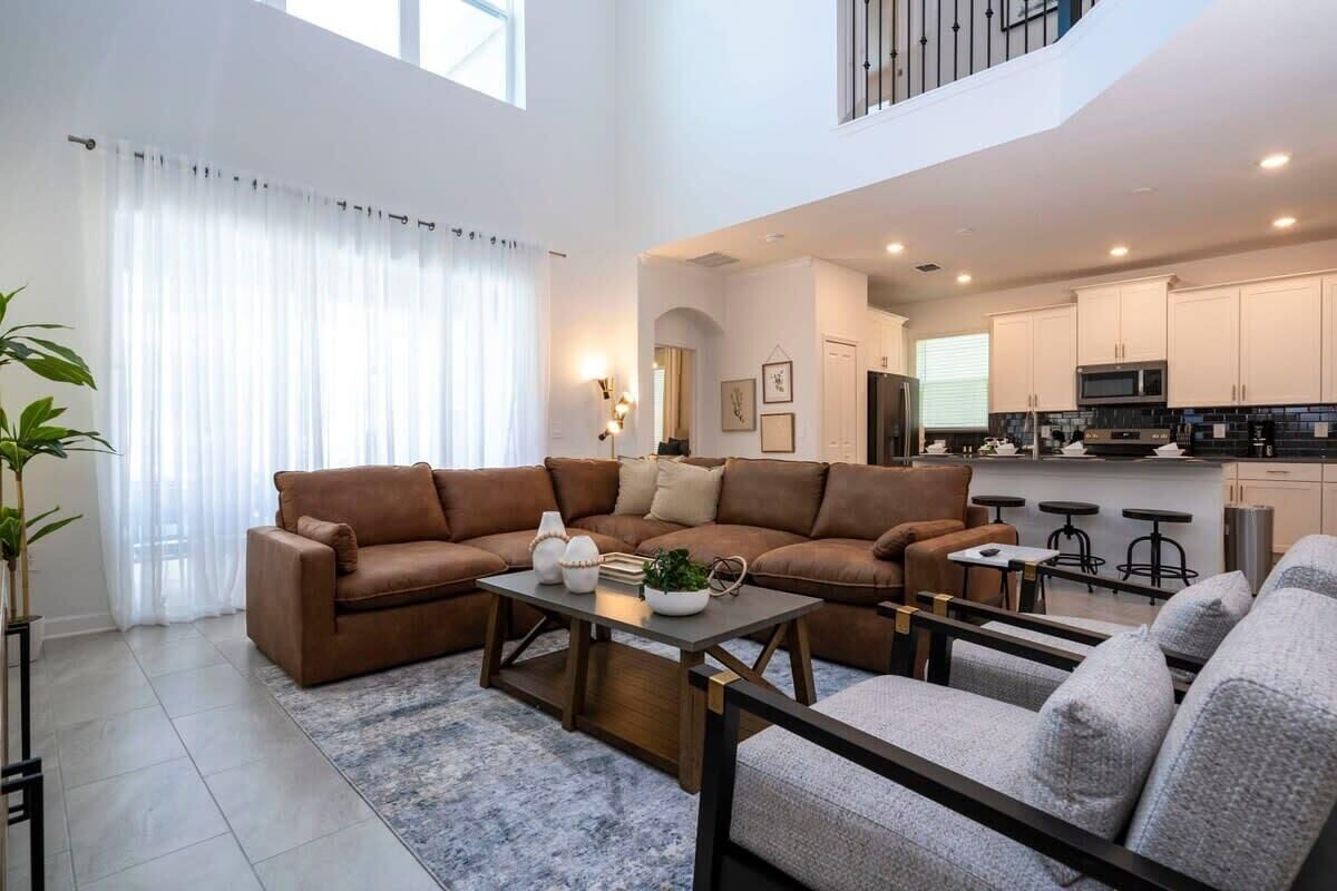 Amplia sala de estar con un sofá seccional marrón, un sillón gris y una mesa de centro. Se observa una cocina al fondo con gabinetes blancos y un microondas.
