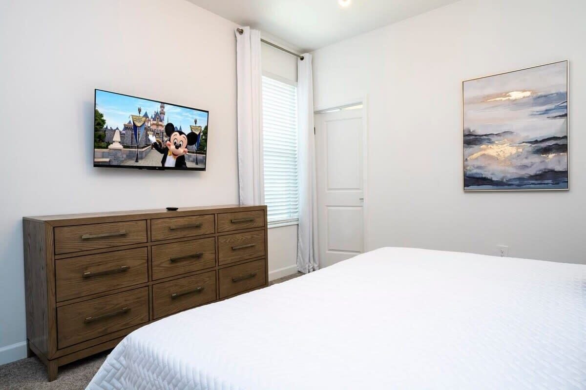A bedroom with a bed, a wooden dresser, a television displaying a cartoon character, and a painting on the wall.