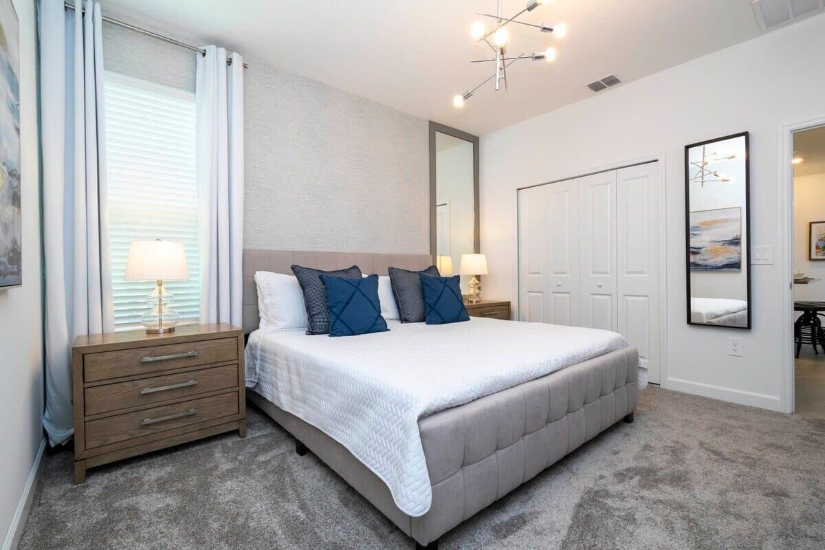 A bedroom with a large bed, a nightstand with a lamp, a dresser, a wardrobe, and a mirror on the wall.