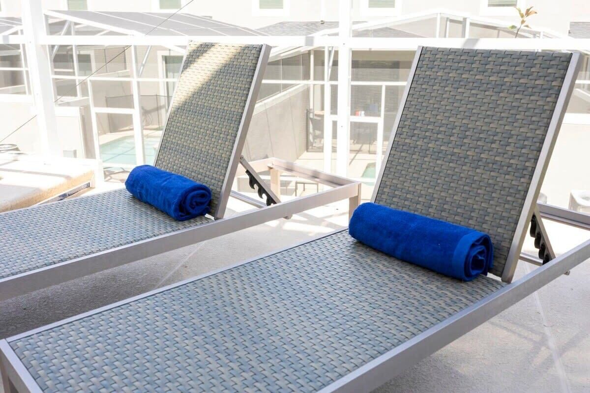 Dos sillones de descanso con cojines azules, estructura metálica y respaldos texturizados, ubicados en un balcón con baranda de vidrio y vistas a una piscina y un edificio.