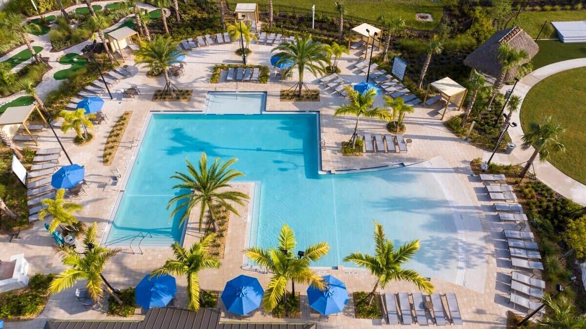 Ein Luftbild zeigt ein Resort mit einem zentralen Schwimmbecken, umgeben von Liegestühlen und Sonnenschirmen, Palmen und einem gepflegten Rasen.