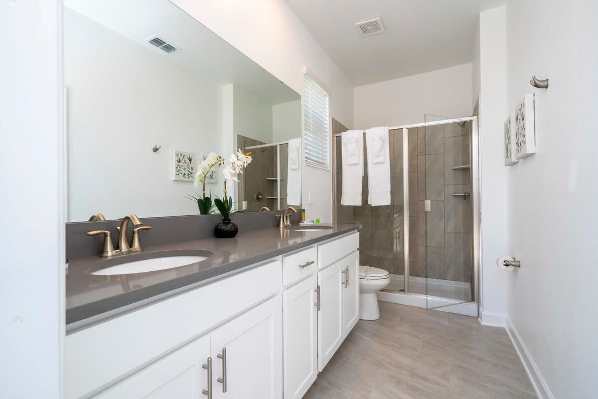 A modern bathroom with a large mirror, two sinks, a shower, and a toilet.