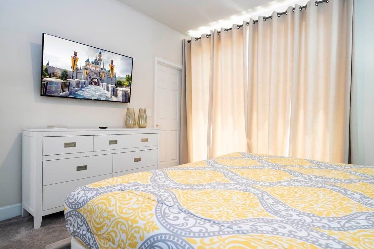 A bedroom with a bed, a TV mounted on the wall, a white dresser, and a window with curtains.