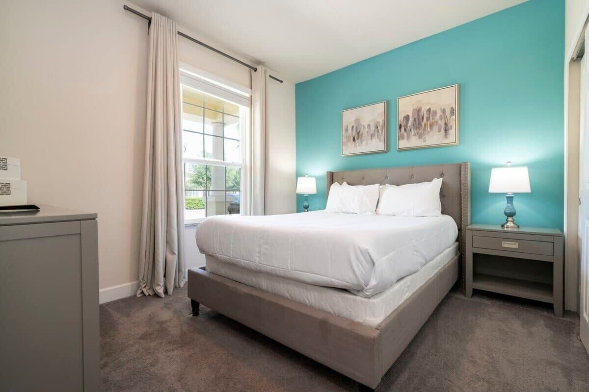 Une chambre à coucher avec un grand lit, deux tables de chevet avec des lampes, une fenêtre avec des rideaux et des œuvres d’art accrochées au mur.