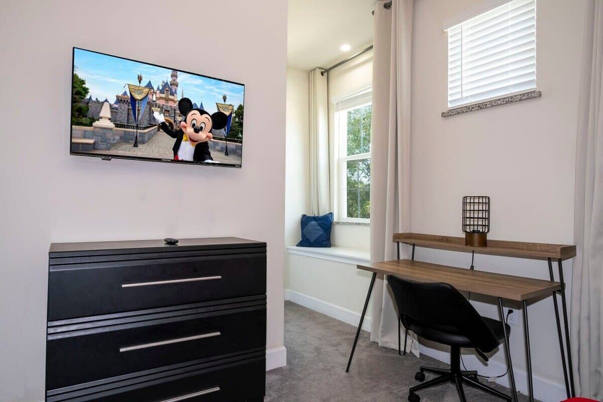 Ein Zimmer mit Fernseher, der Mickey Mouse zeigt, einer schwarzen Kommode, einem Holztisch, einem schwarzen Bürostuhl und einem Fenster mit Blick auf Bäume.