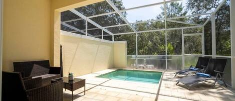 A sunlit patio with a pool, wicker furniture, and a glass enclosure.
