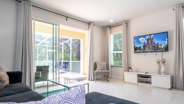 A modern living room with a sofa, a TV mounted on the wall, and a balcony with glass doors. - Marvelous 3Bd w/ Pool Near Disney at Serenity 1536 (Clermont)