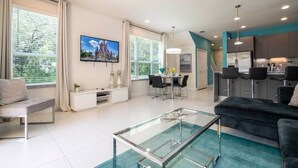 A modern living room with a glass coffee table, a black sofa, and a dining area with a TV displaying a castle. - Marvelous 3Bd w/ Pool Near Disney at Serenity 1536 (Clermont)