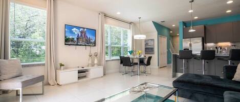 A modern living room with a glass coffee table, a black sofa, and a dining area with a TV displaying a castle.
