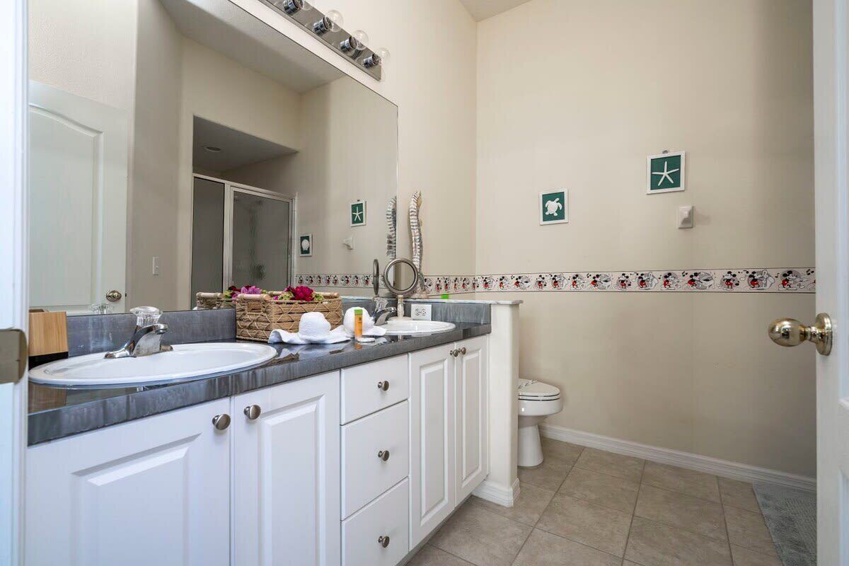 Casa de banho com lavatório duplo, espelho, sanita e chuveiro. Apresenta azulejos decorativos na parede e um porta-toalhas.