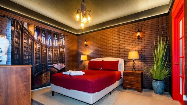 A bedroom with a brick wall, a large bed with red bedding, two bedside lamps, a nightstand, a potted plant, and a red door. - Marvelous 4Bd Close to Disney w/ Pool at 313 Tuscan Hills (Davenport)