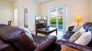A living room with a brown leather sofa, a wooden coffee table, and a TV on a stand. There is a sliding glass door leading to a pool area. - Marvelous 4Bd Close to Disney w/ Pool at 313 Tuscan Hills (Davenport)