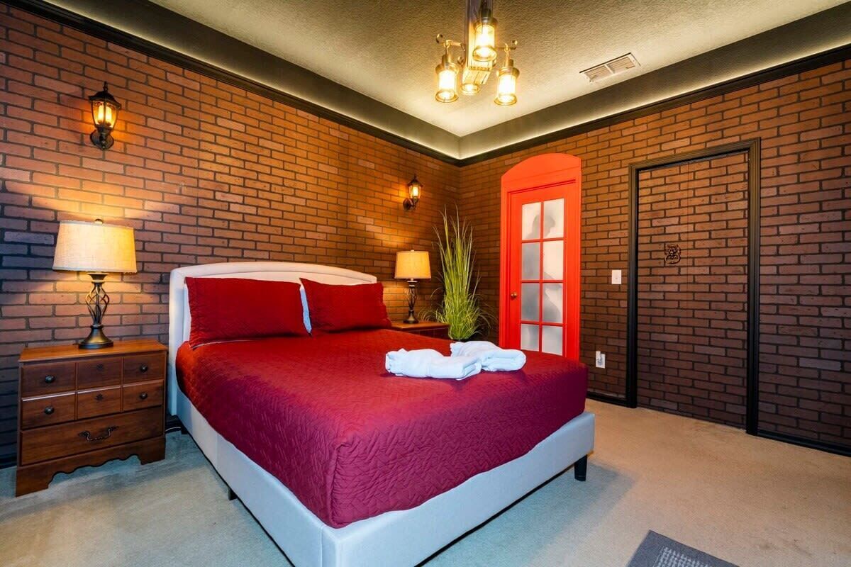A bedroom with a brick wall, a red door, a bed with red bedding, two bedside tables with lamps, and a chandelier.