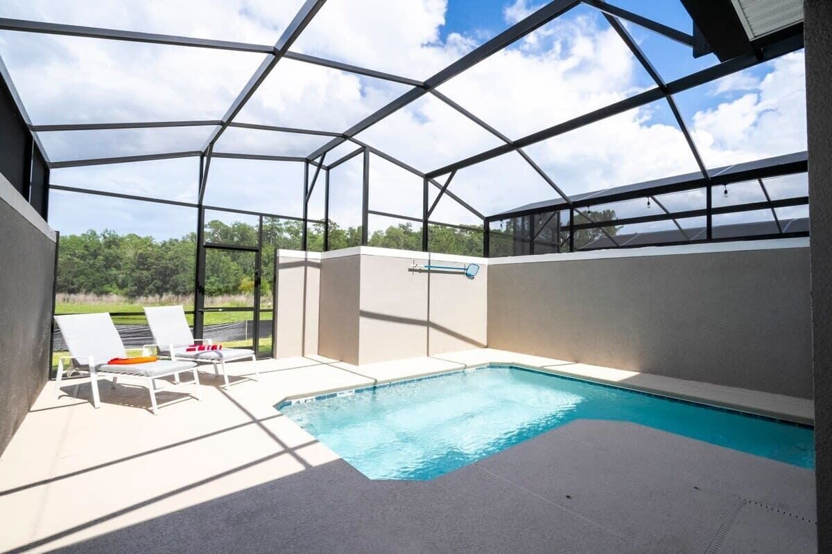 An indoor swimming pool with a glass enclosure, two lounge chairs, and a view of the outdoors.