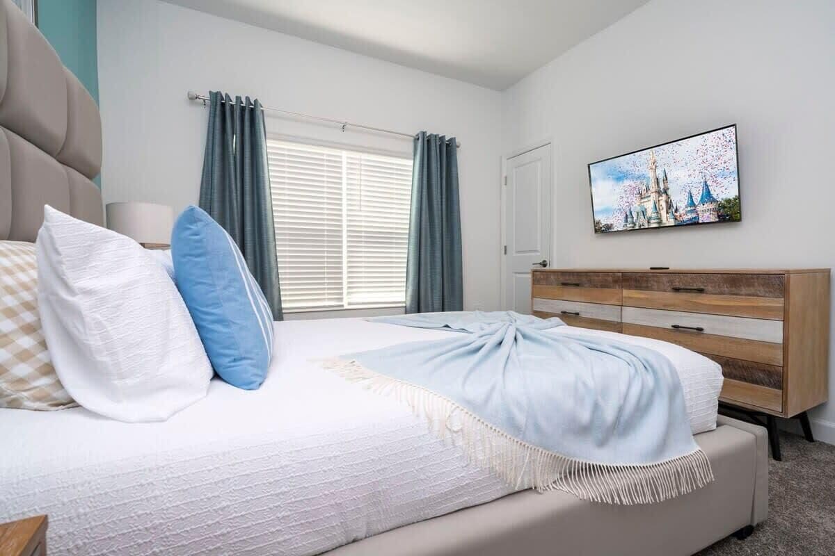 Una camera da letto con un letto, una cassettiera, una televisione a parete e una finestra con tende.