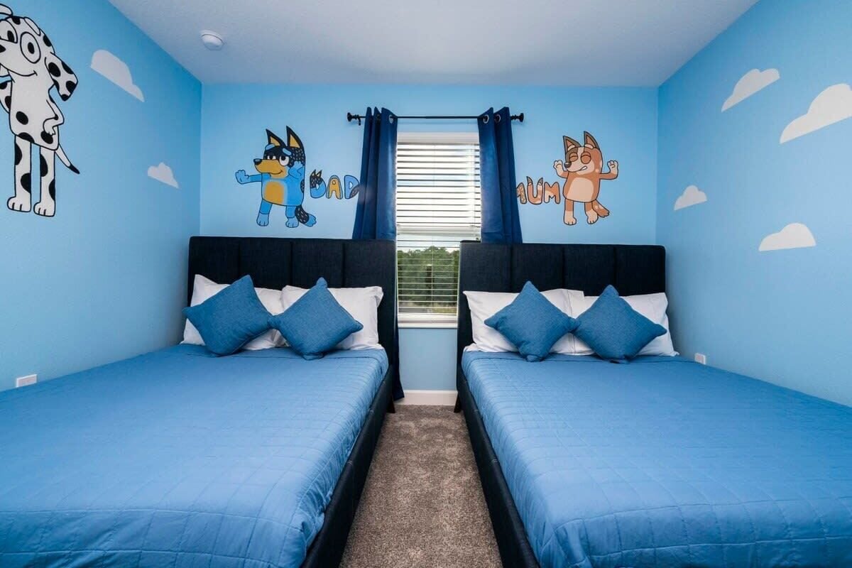Ein Zimmer mit zwei Betten, jeweils mit blauer Bettwäsche und weißen Kissen, einem Fenster mit blauen Vorhängen sowie Wanddekorationen, darunter Zeichentrickfiguren.