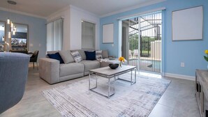A modern living room with a sofa, coffee table, and a view of the outdoors through large windows. - Marvelous 4Bd with Pool at Storey Lake Resort 2601 (Kissimmee)