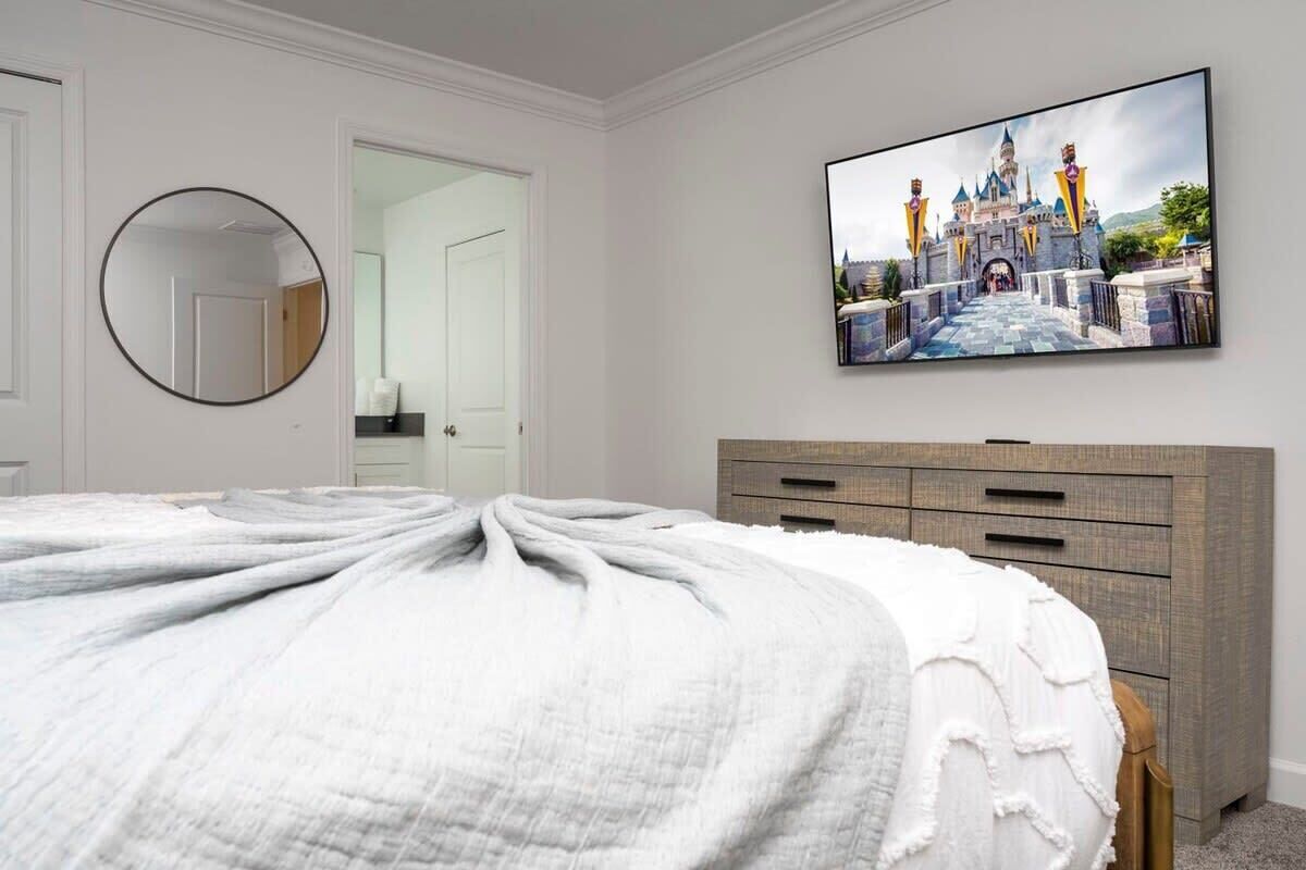 Une chambre à coucher avec un grand lit, une commode, un miroir et un téléviseur affichant une image de château.