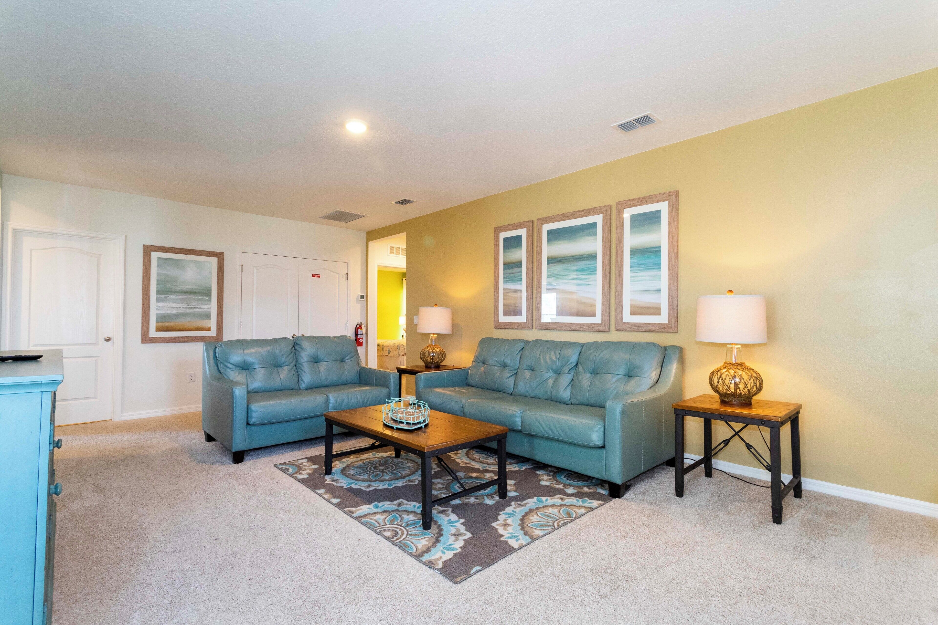 A living room with two blue leather sofas, a wooden coffee table, and a patterned rug. There are three framed pictures on the wall and two lamps on side tables.
