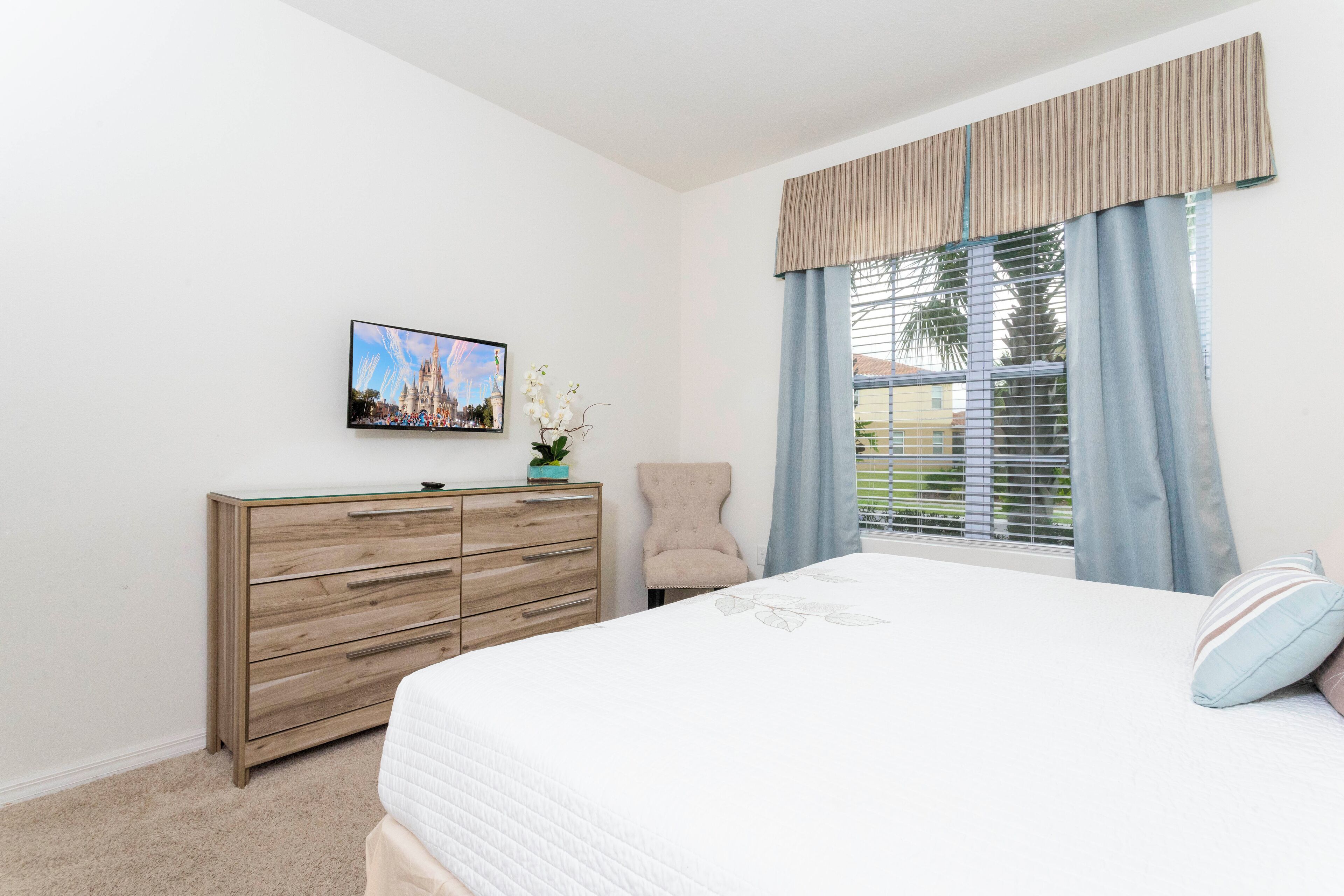 A bedroom with a bed, a wooden dresser, a chair, a television on the wall, and a window with curtains.