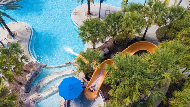 Luchtbeeld van een zwembadgedeelte van een resort, met een waterglijbaan, palmbomen en een helderblauw zwembad.
