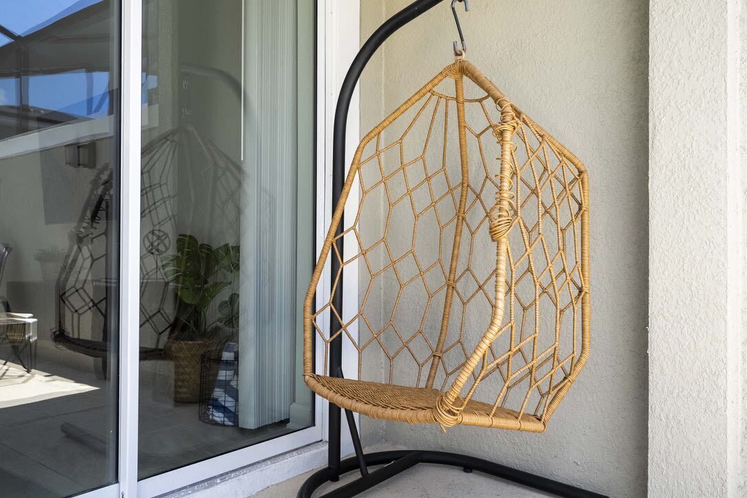 Una silla colgante de mimbre con diseño geométrico, sujeta a un soporte de metal negro, ubicada frente a una pared con puerta de vidrio que refleja una planta y una silla.