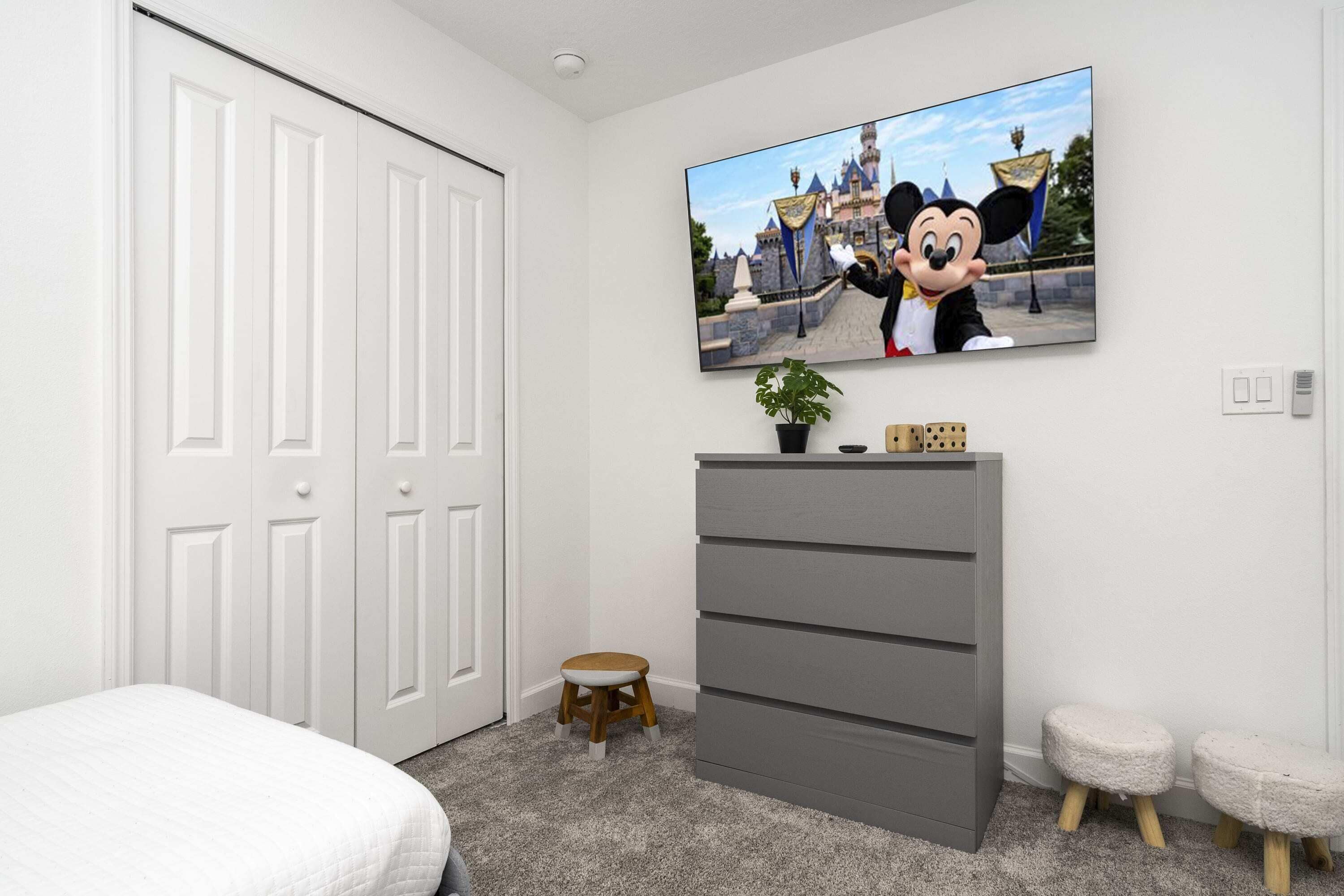 Ein Schlafzimmer mit einem Bett, einer grauen Kommode, einem Fernseher, der einen Zeichentrickcharakter zeigt, und einem kleinen Hocker.