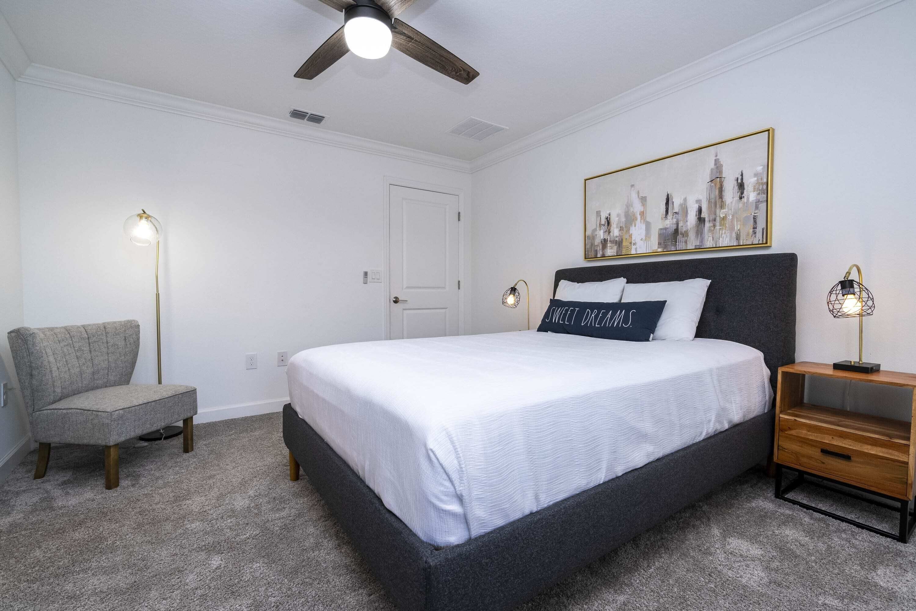 Une chambre à coucher comprenant un lit, une chaise, une table de chevet, un ventilateur de plafond et une œuvre d’art accrochée au mur.