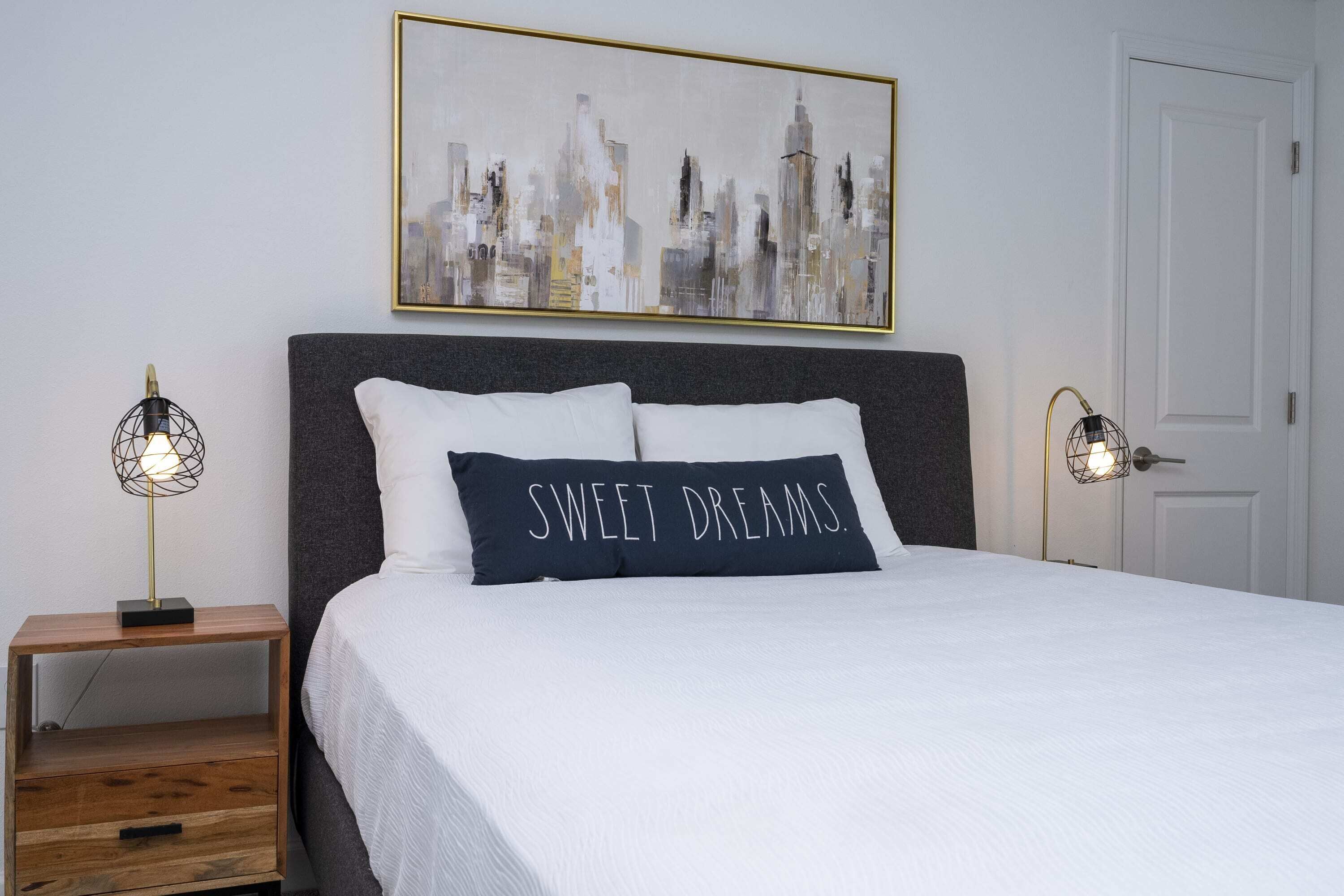 A neatly made bed with a pillow that reads "SWEET DREAMS." A cityscape painting hangs above the bed, and there are two bedside lamps on either side.
