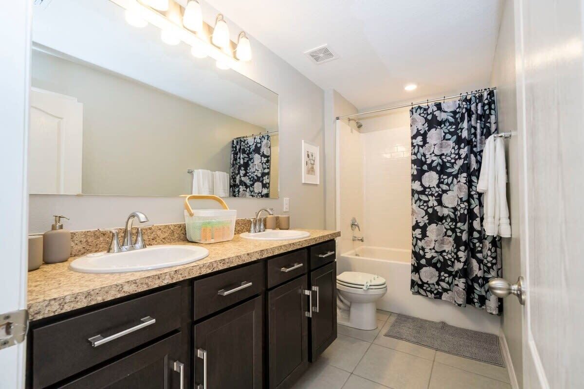 A bathroom with two sinks, a large mirror, a shower with floral curtains, and a toilet.