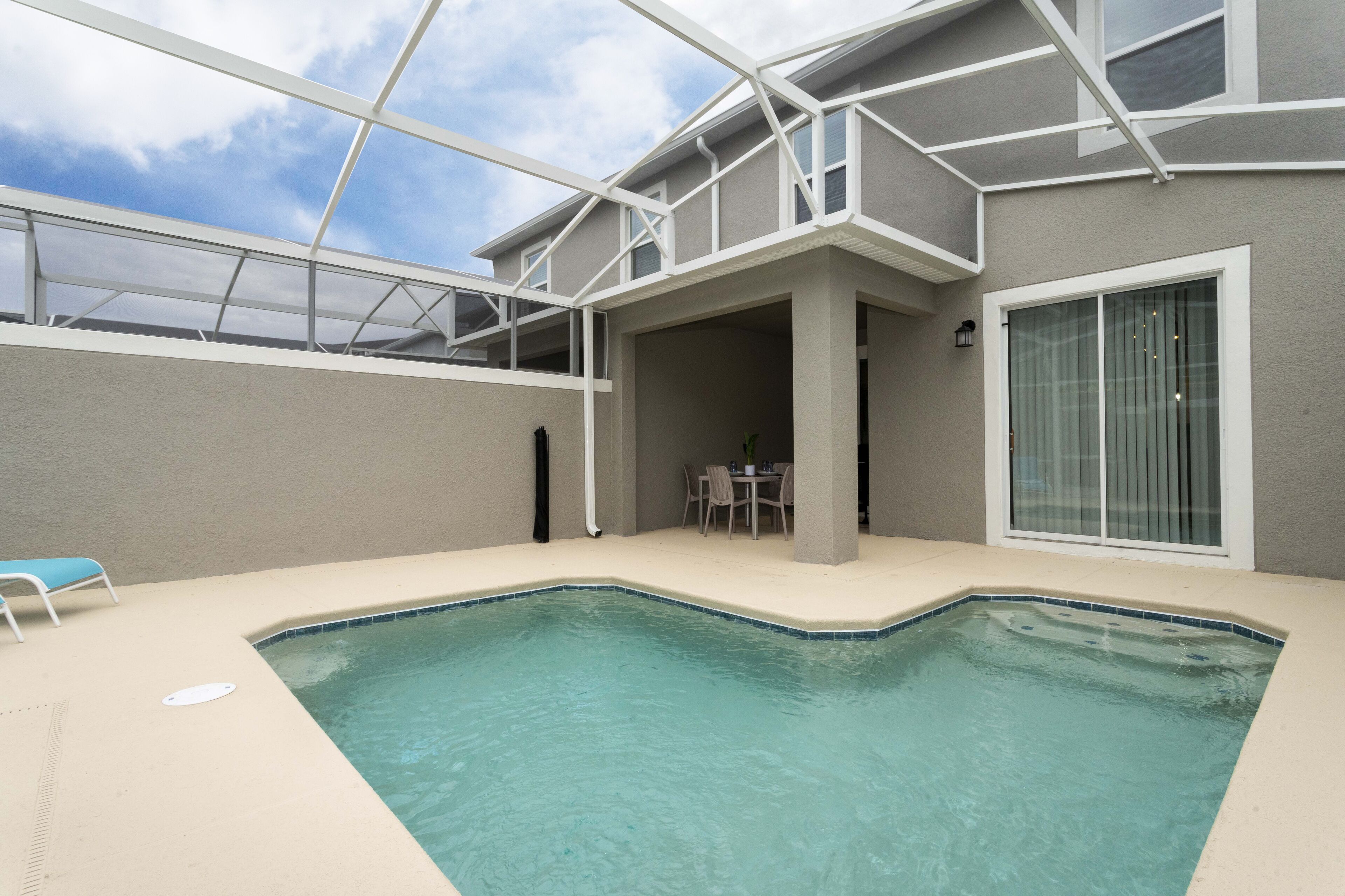 A swimming pool with a covered patio area, a table and chairs, and a large window.