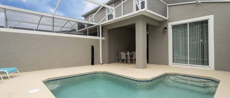 A swimming pool with a covered patio area, a table and chairs, and a large window.