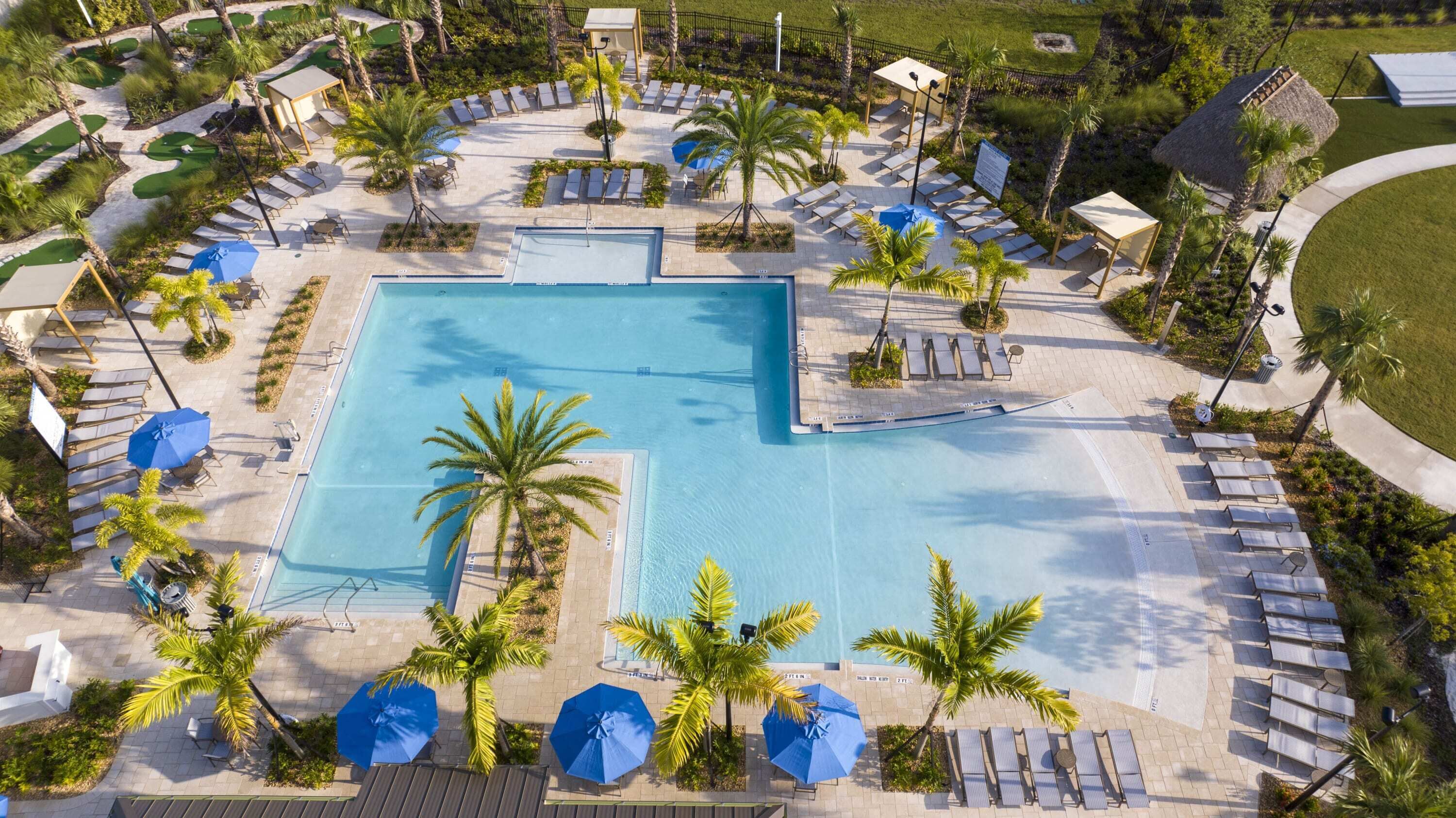 Una gran piscina rodeada de sillones de descanso y sombrillas, con palmeras y un campo de golf al fondo.
