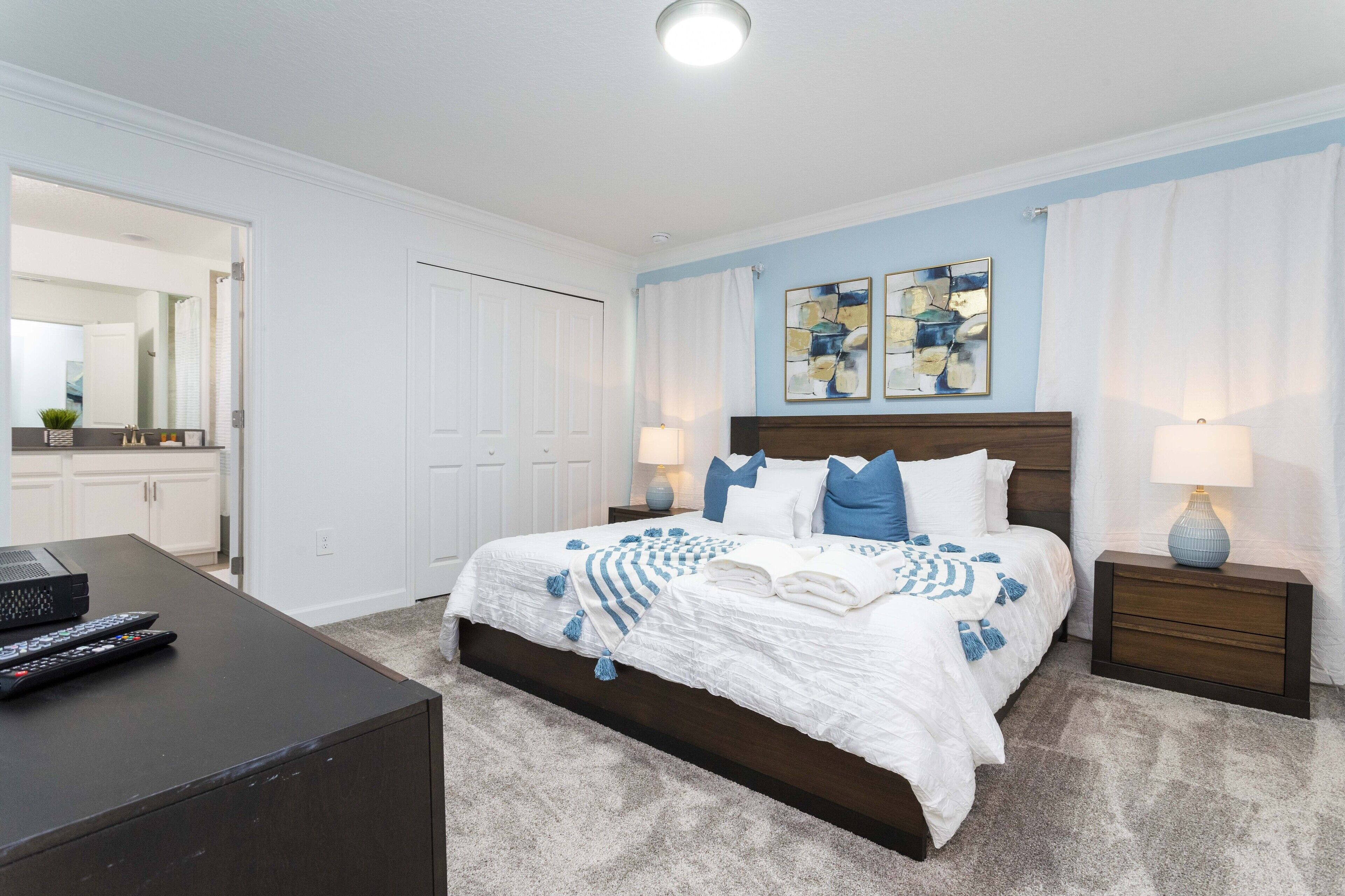 A bedroom with a large bed, two bedside tables with lamps, a dresser, and a desk with a computer and remote controls.