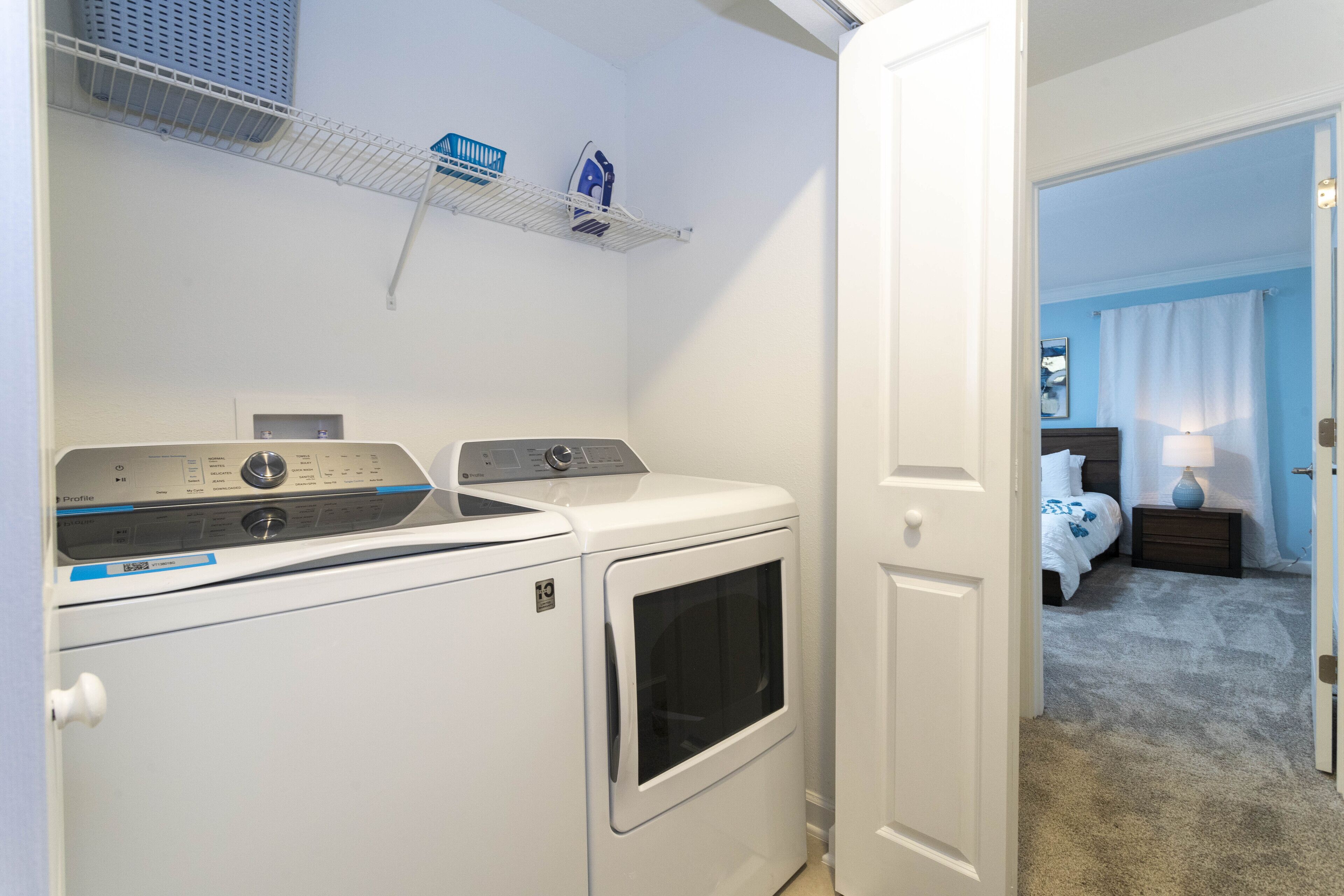 A laundry room with a front-loading washer and dryer, a shelf with laundry supplies, and a bedroom visible through an open door.