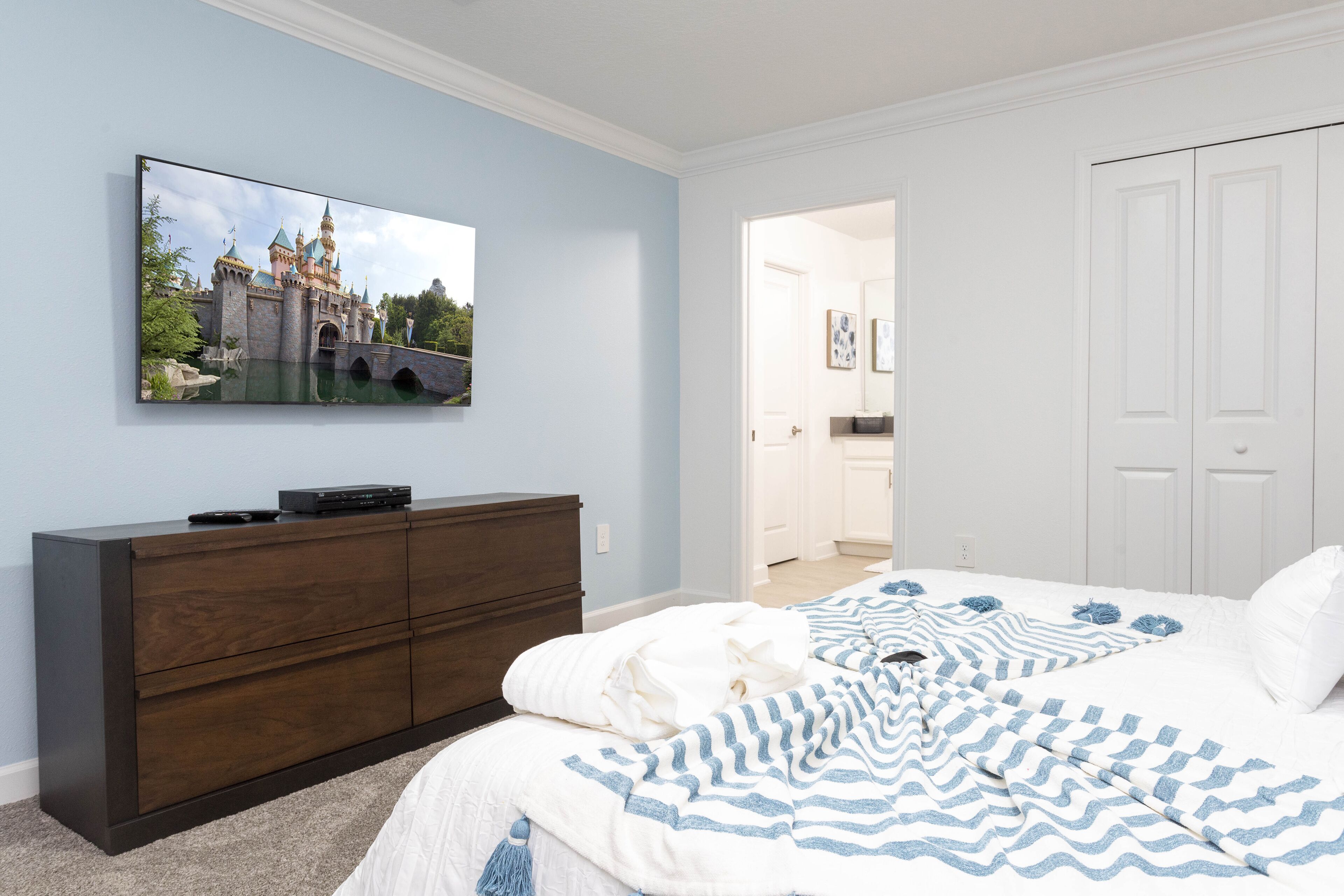 Une chambre à coucher comprenant un lit, une commode, un téléviseur fixé au mur et une porte donnant sur une autre pièce.