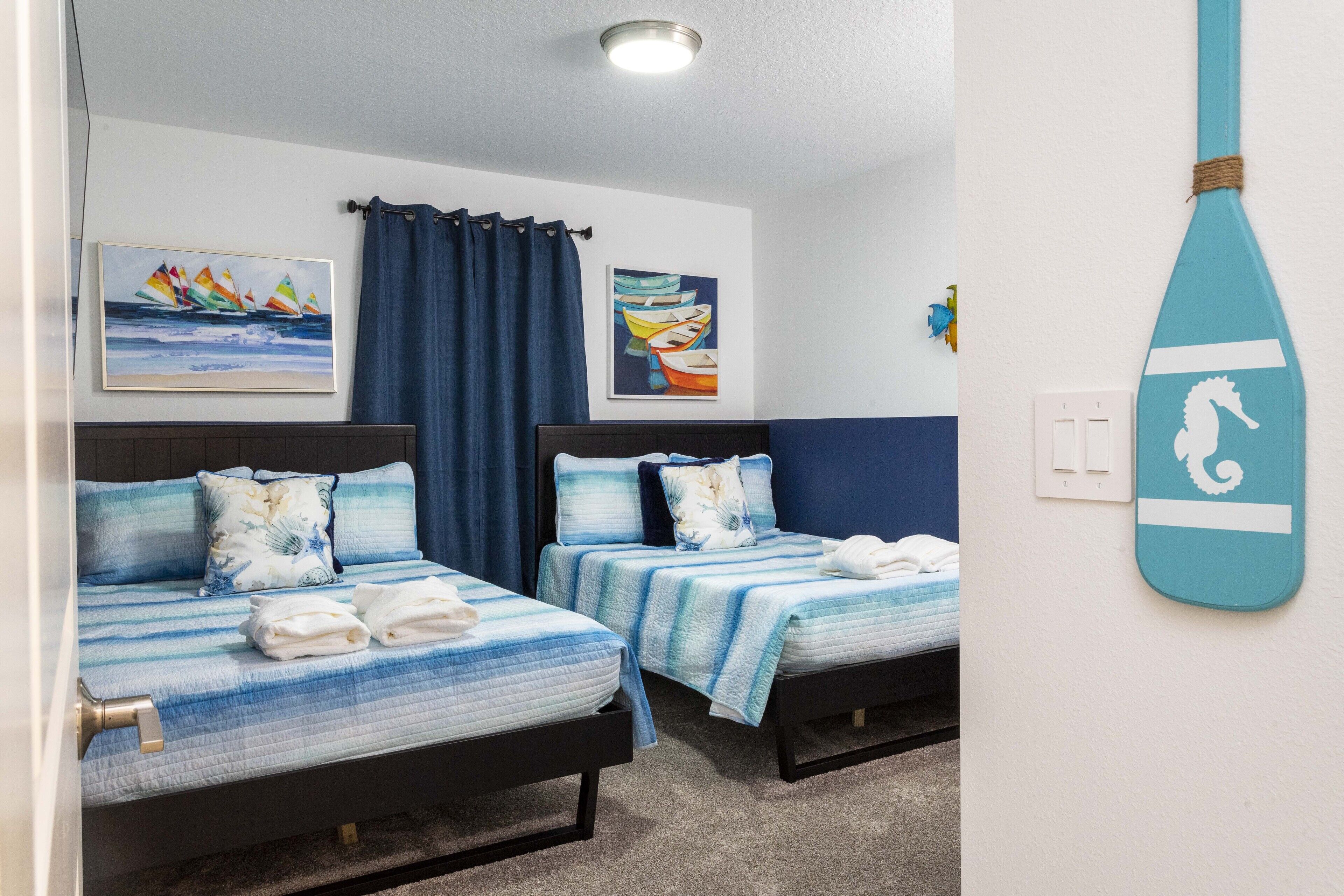 A hotel room with two beds, each with matching blue bedding and pillows, a nautical-themed wall decoration, and a blue curtain.