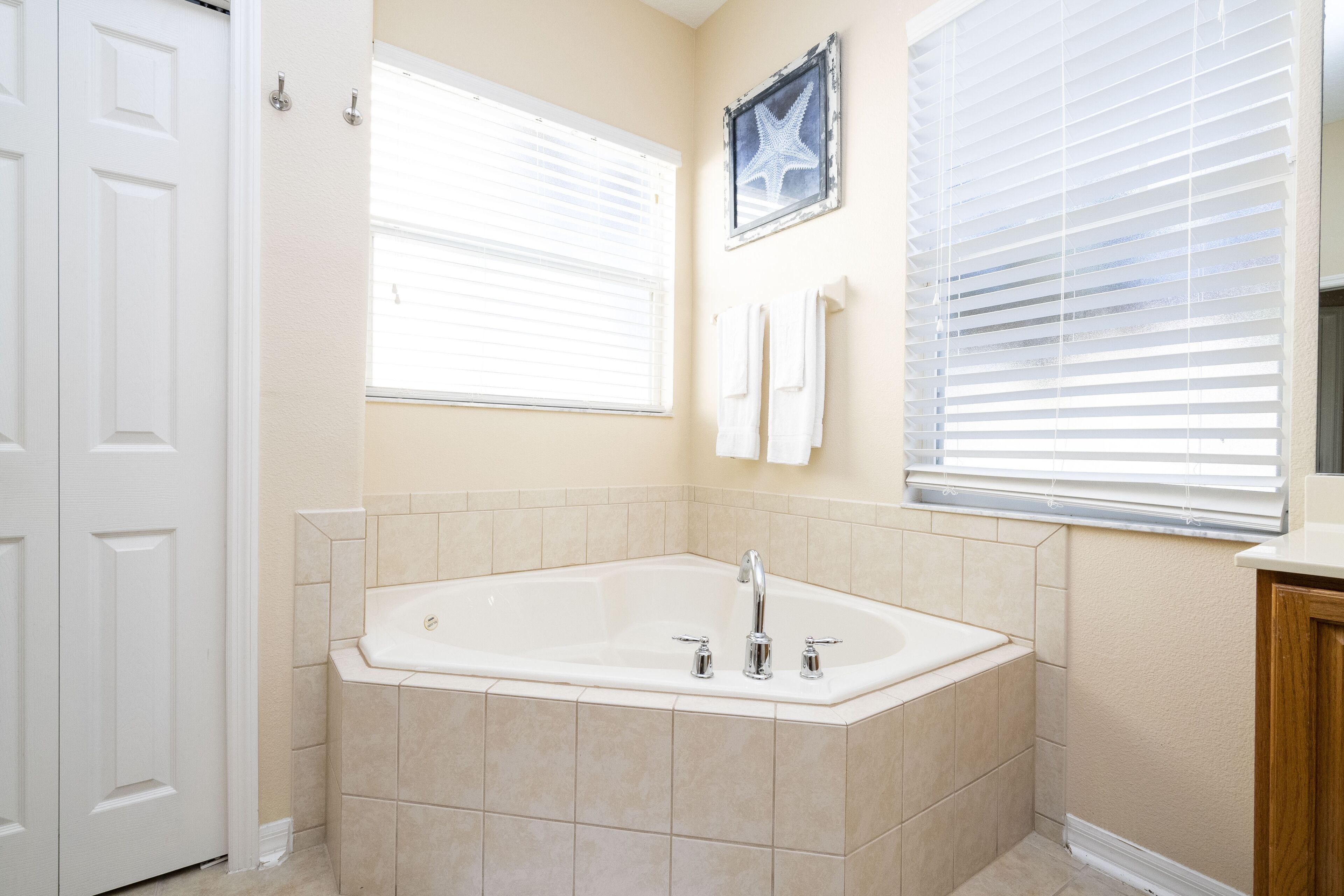 Ein Badezimmer mit einer großen Badewanne, beige gefliesten Wänden und einem Fenster mit Jalousien.