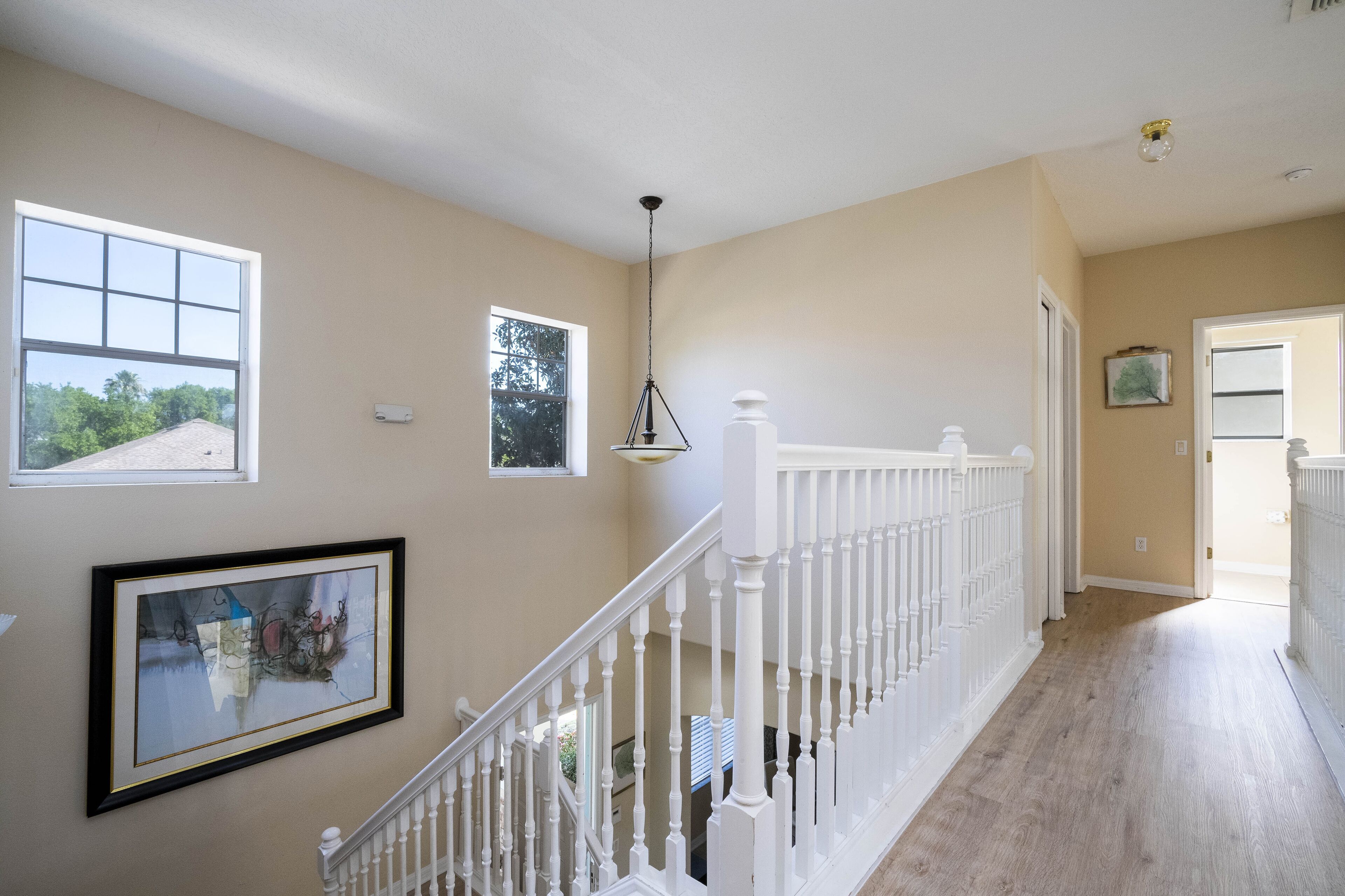 A spacious hallway with a wooden floor, white railing, and multiple windows offering views of the outdoors.