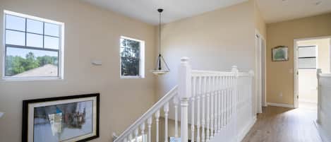 A spacious hallway with a wooden floor, white railing, and multiple windows offering views of the outdoors.