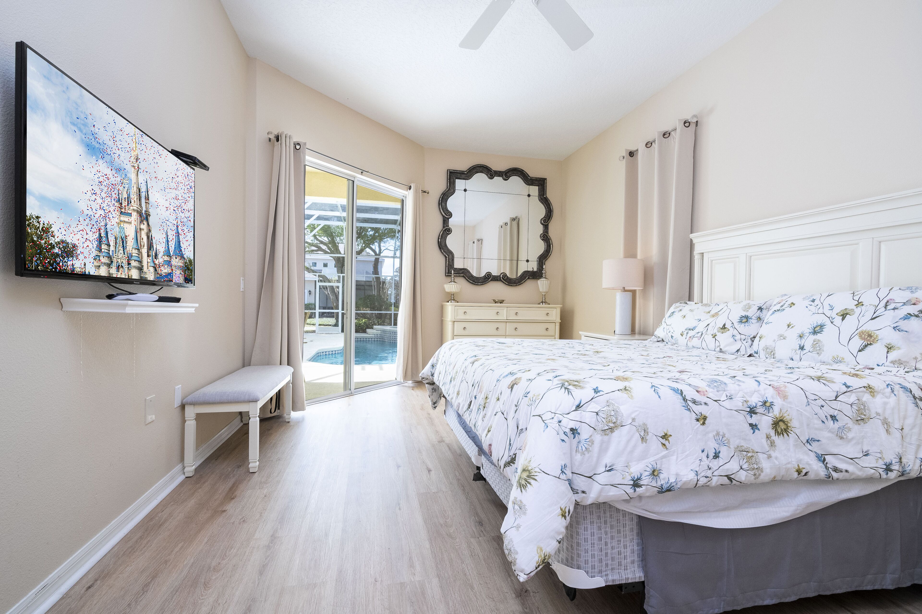 Ein Schlafzimmer mit einem großen Bett, einem an der Wand montierten Fernseher, einer Bank, einem Schminktisch und einer Schiebetür, die auf einen Balkon führt.