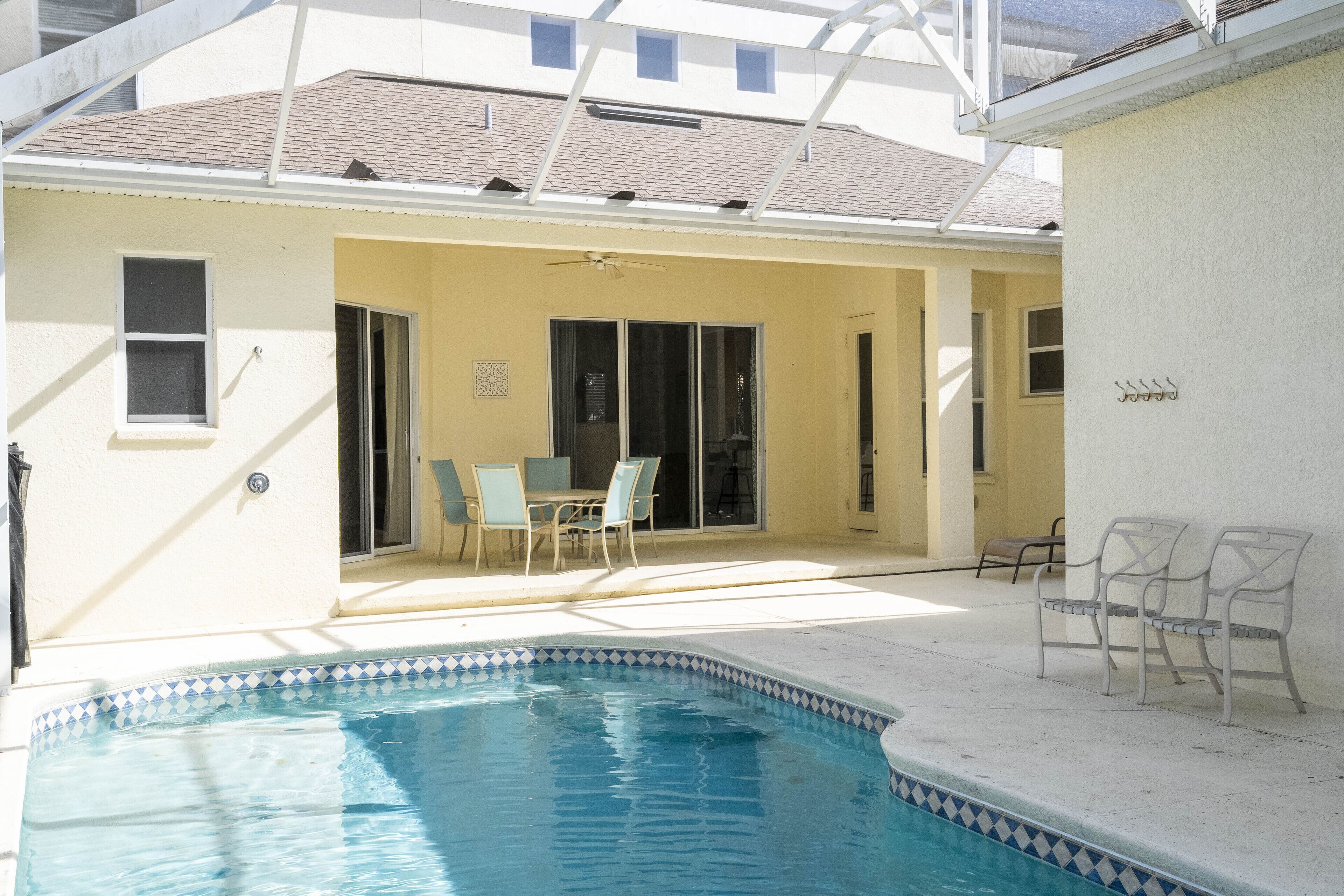 A swimming pool with a tiled edge, a covered patio area with chairs, and a building with a flat roof and windows.