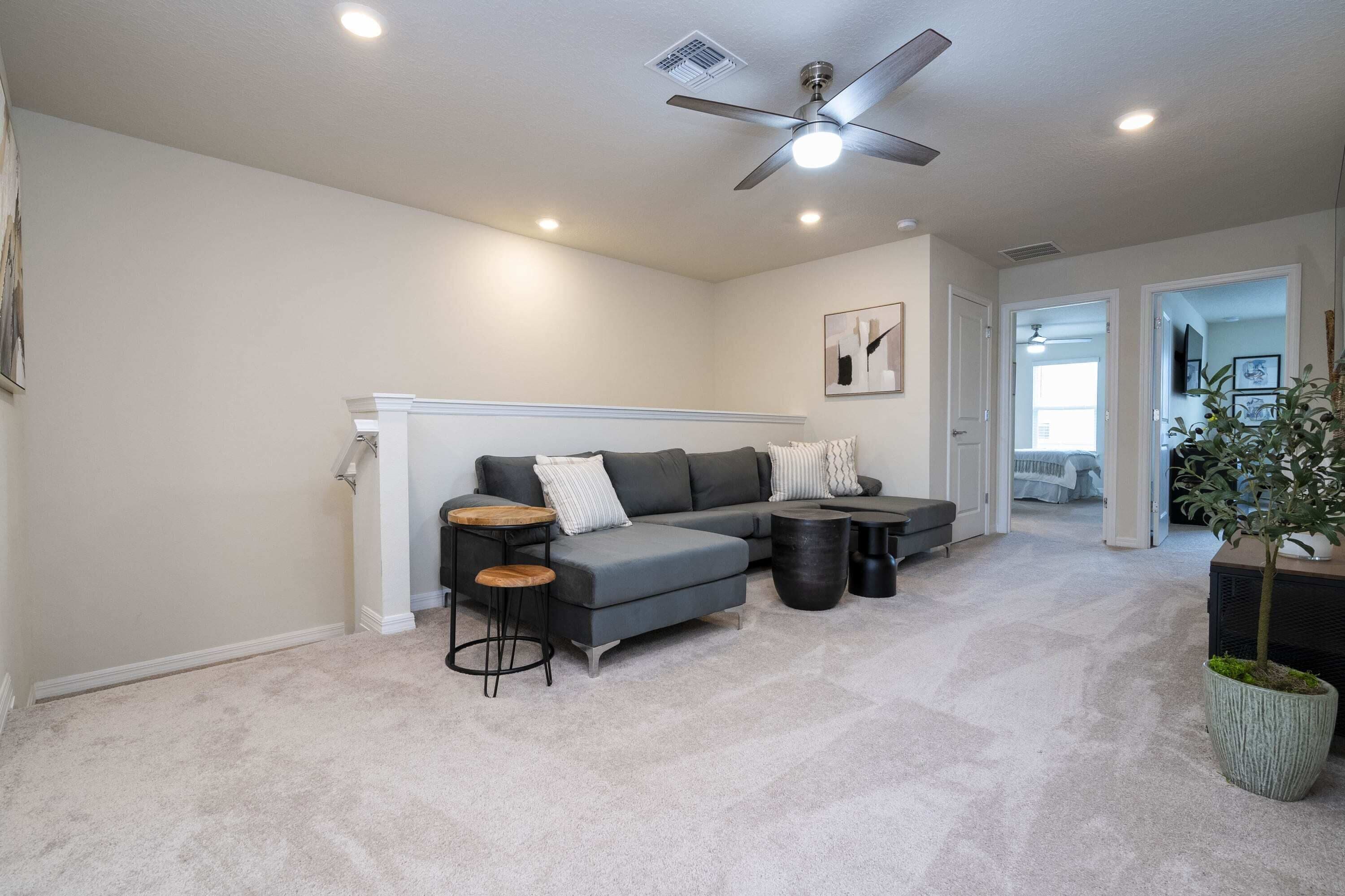 Un salon moderne avec un canapé d’angle gris, un ventilateur de plafond et une grande plante en pot.