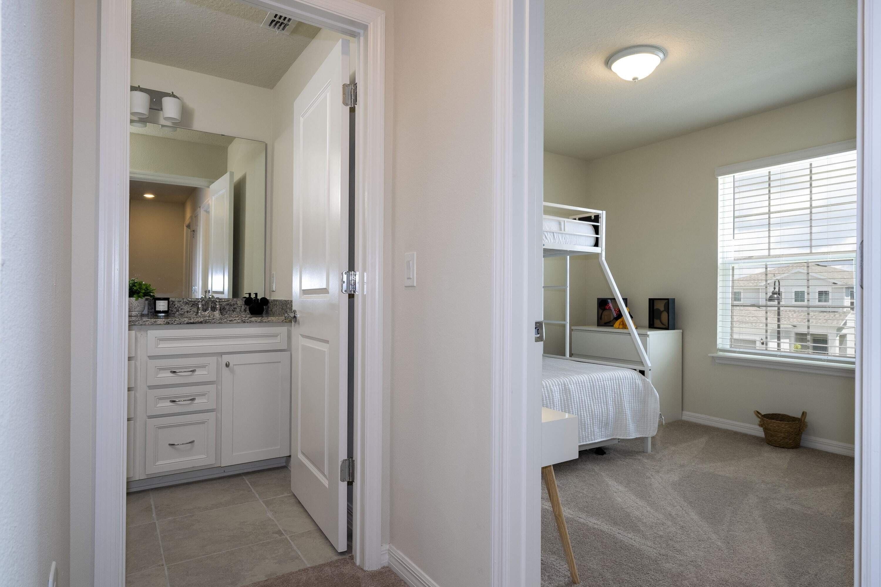 A bedroom with a bunk bed, a large window with blinds, a bedside table, and a carpeted floor.