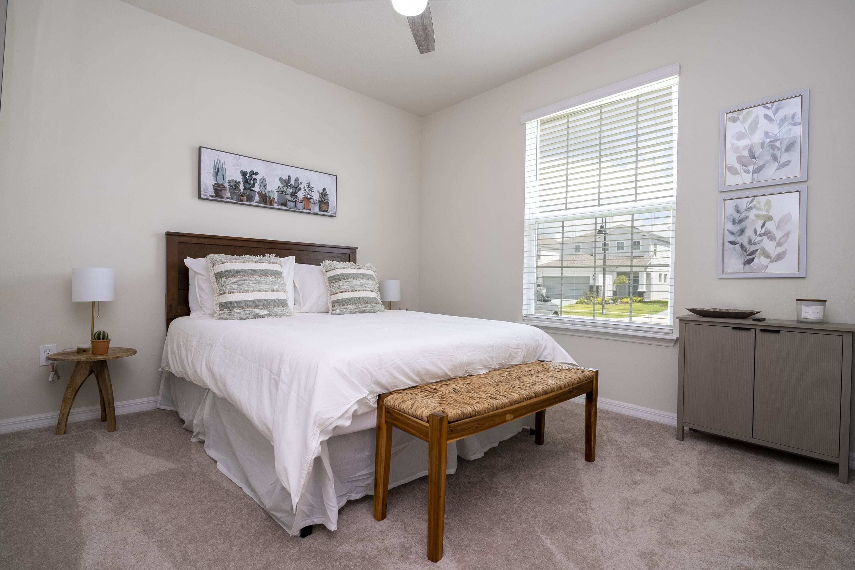 A bedroom with a bed, bedside table, lamp, window, and artwork.