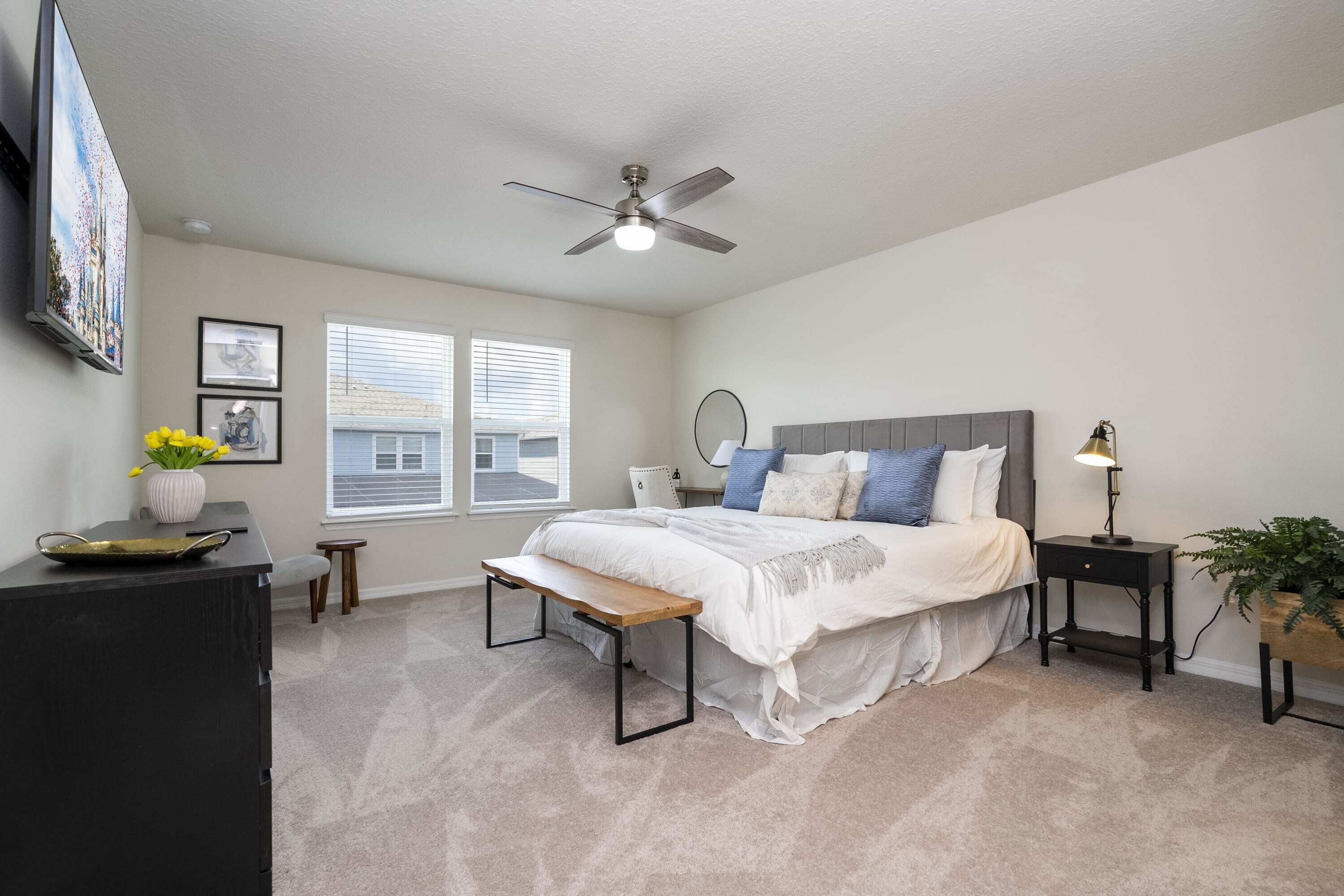 A bedroom with a bed, bedside tables, a ceiling fan, a window, and a desk with a plant.